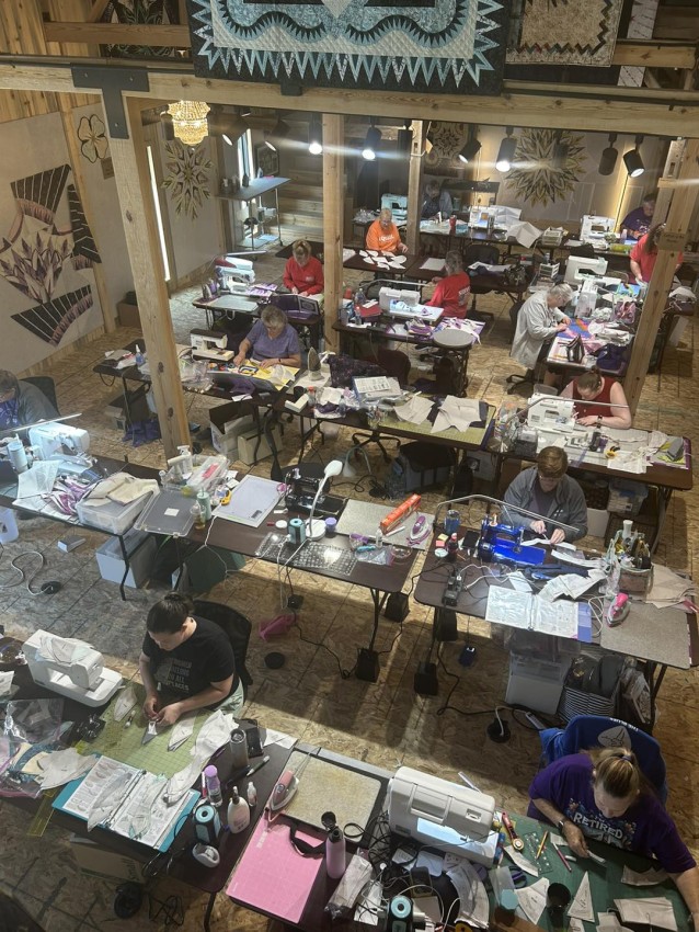 Quilters at multiple tables working on a project with an instructor at the front.